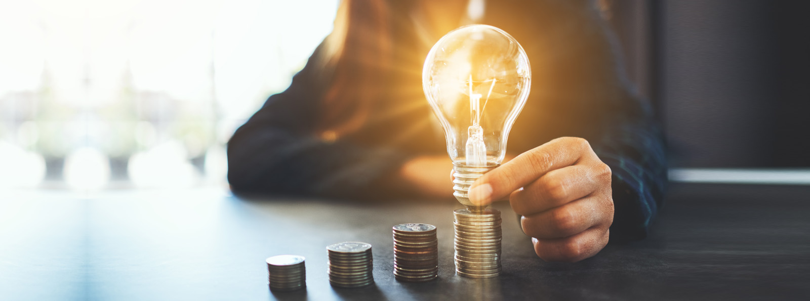 business person holding lit lightbulb over stacks of coins