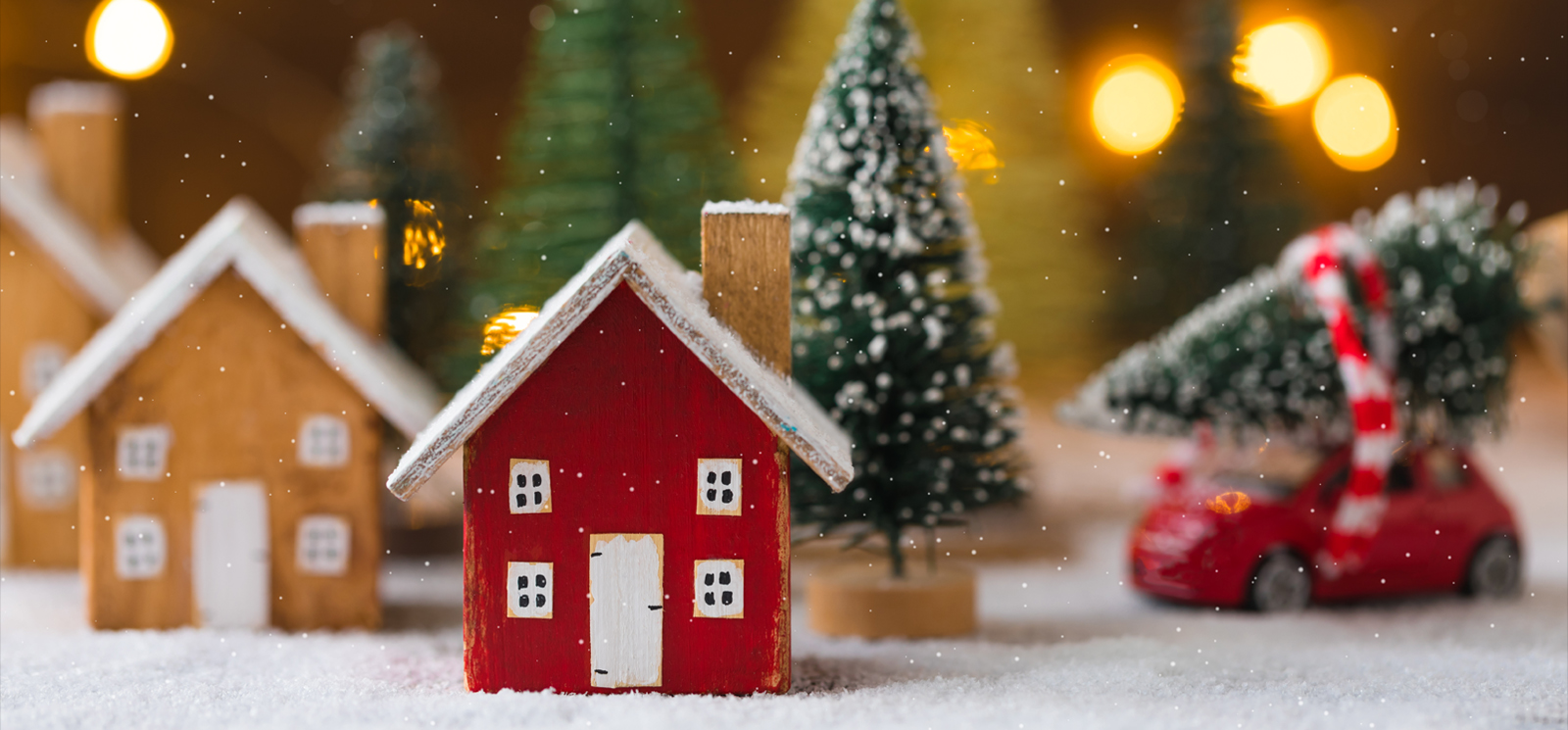 Miniature wooden houses and small red car with fir tree on the snow over blurred Christmas decoration background